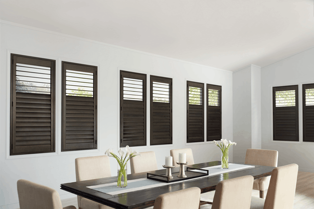 Dark wood plantation shutters with angled tilt louvers installed in a sunlit dining room, offering light control and a warm, natural aesthetic